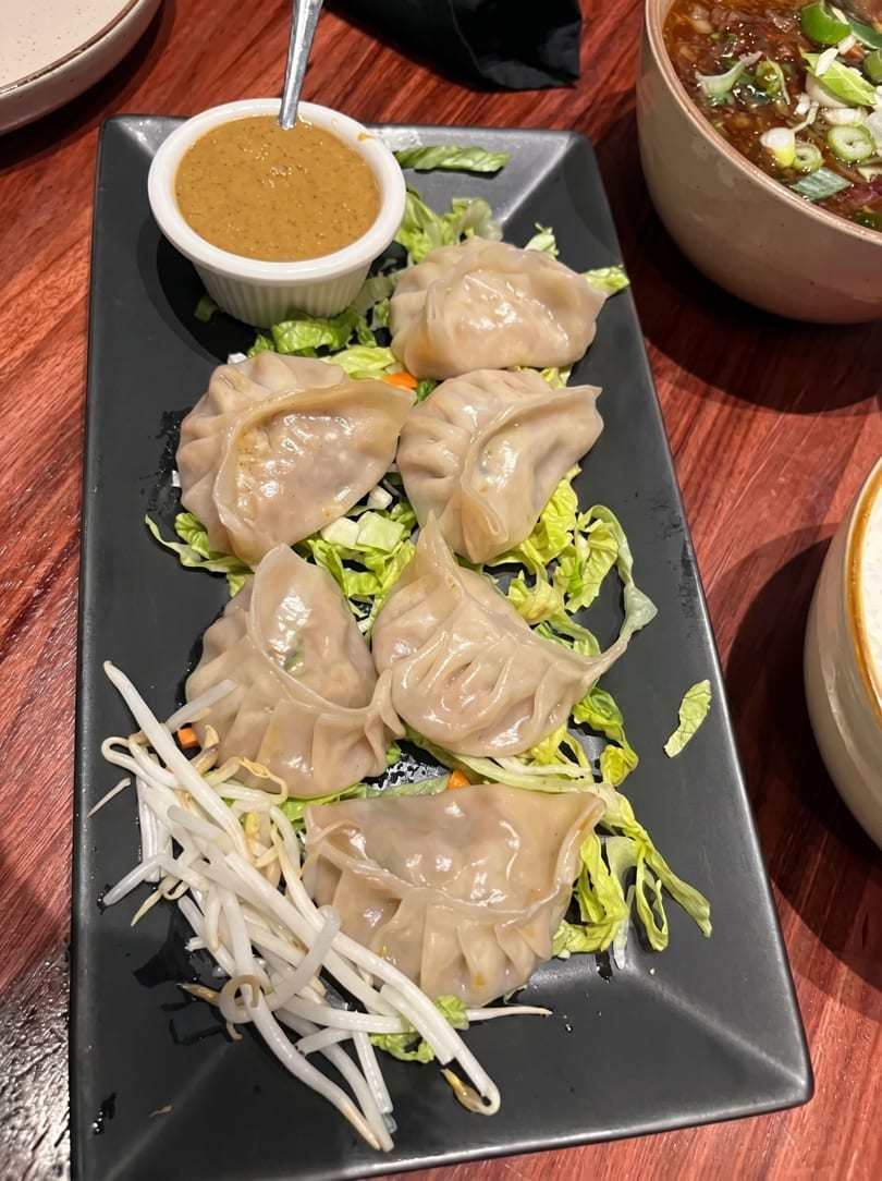 A black rectangular plate of steamed dumplings on lettuce with bean sprouts and a side of peanut sauce.