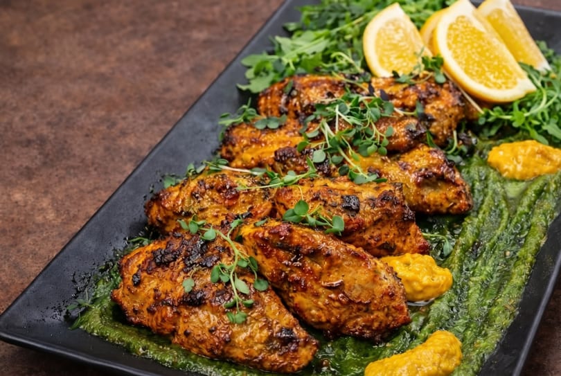 Grilled chicken pieces with green sauce, orange slices, and microgreens on a black plate.