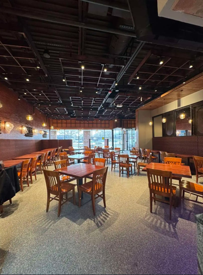 Spacious restaurant interior with numerous empty wooden tables and chairs, brick walls, and large windows.