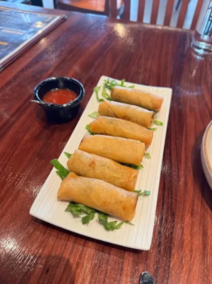 Six crispy spring rolls on a white rectangular plate with a side of red dipping sauce.