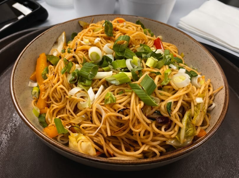 Close-up of a bowl of stir-fried noodles with vegetables and scallions on a dark tray.