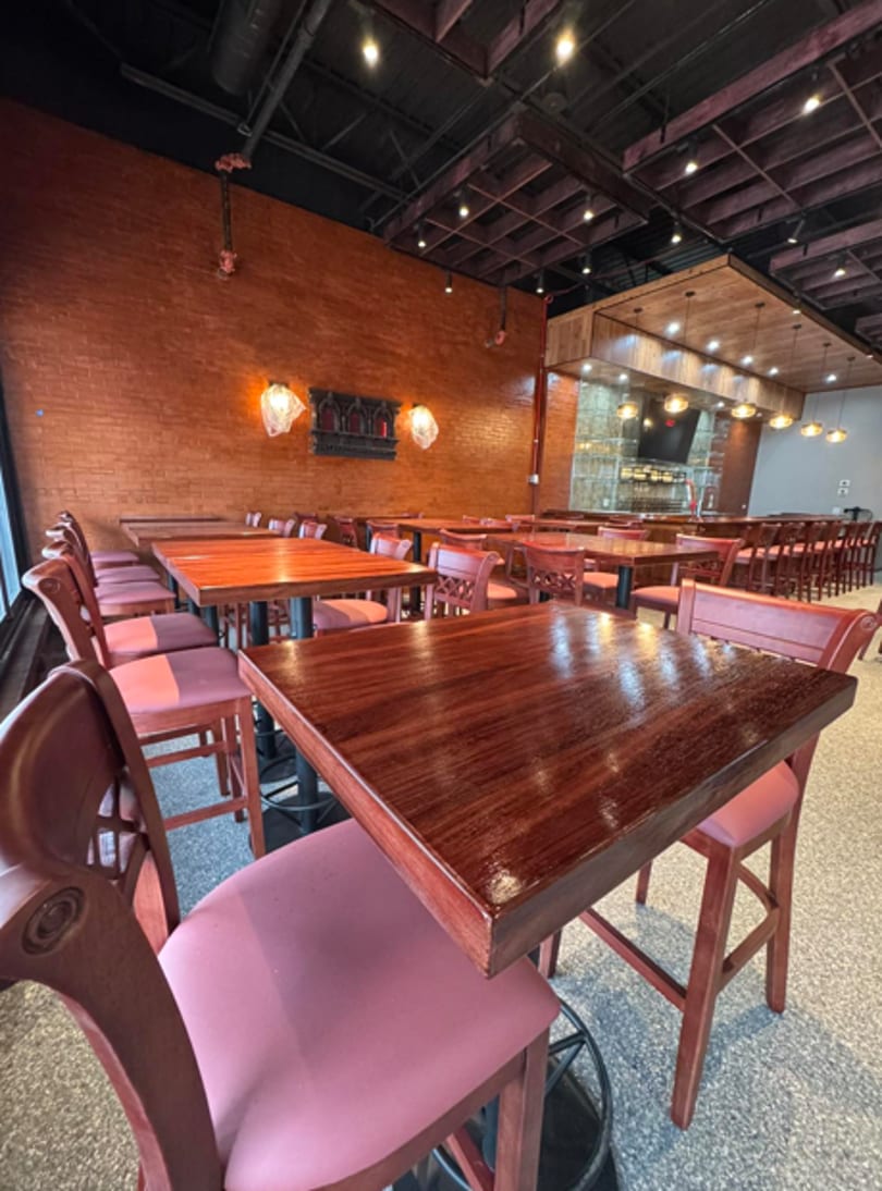 Empty restaurant dining room with dark wooden tables, red chairs, and a brick wall with decorative lighting.