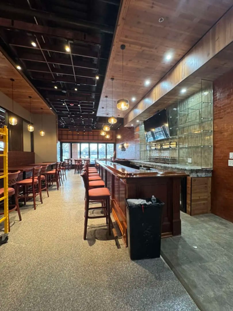 Empty restaurant interior with a long wooden bar, bar stools, and dining tables under warm lighting.