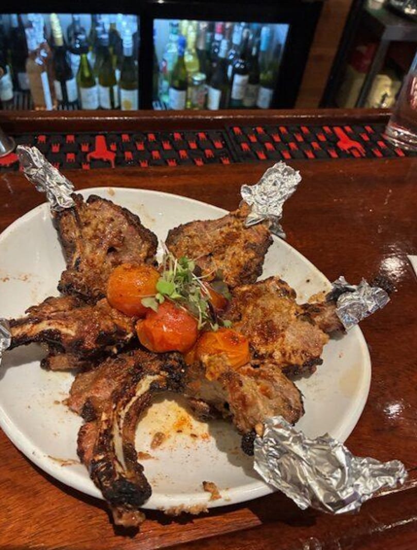 Grilled lamb chops with roasted tomatoes and microgreens on a white plate at a bar.